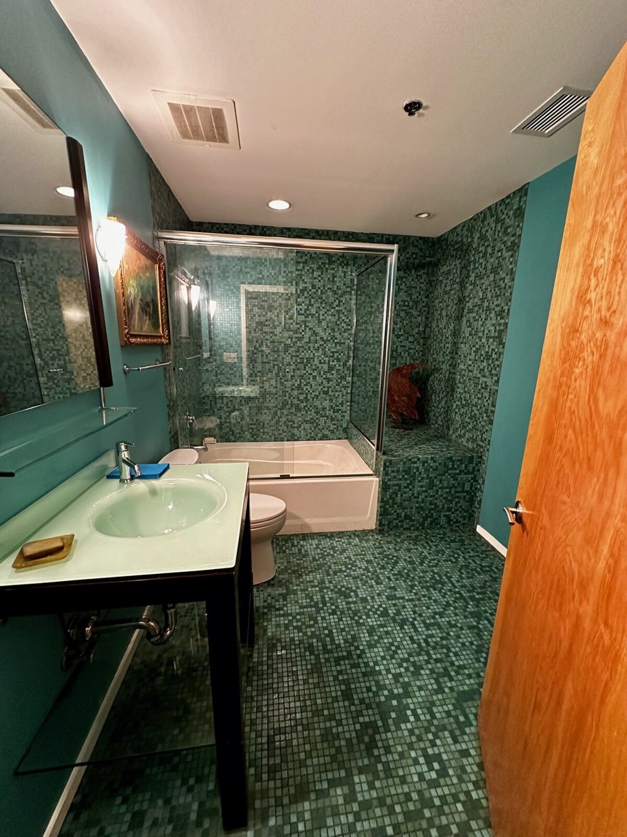 Bathroom before renovation, with green mosaic tile, green walls and a framed sliding tub enclosure